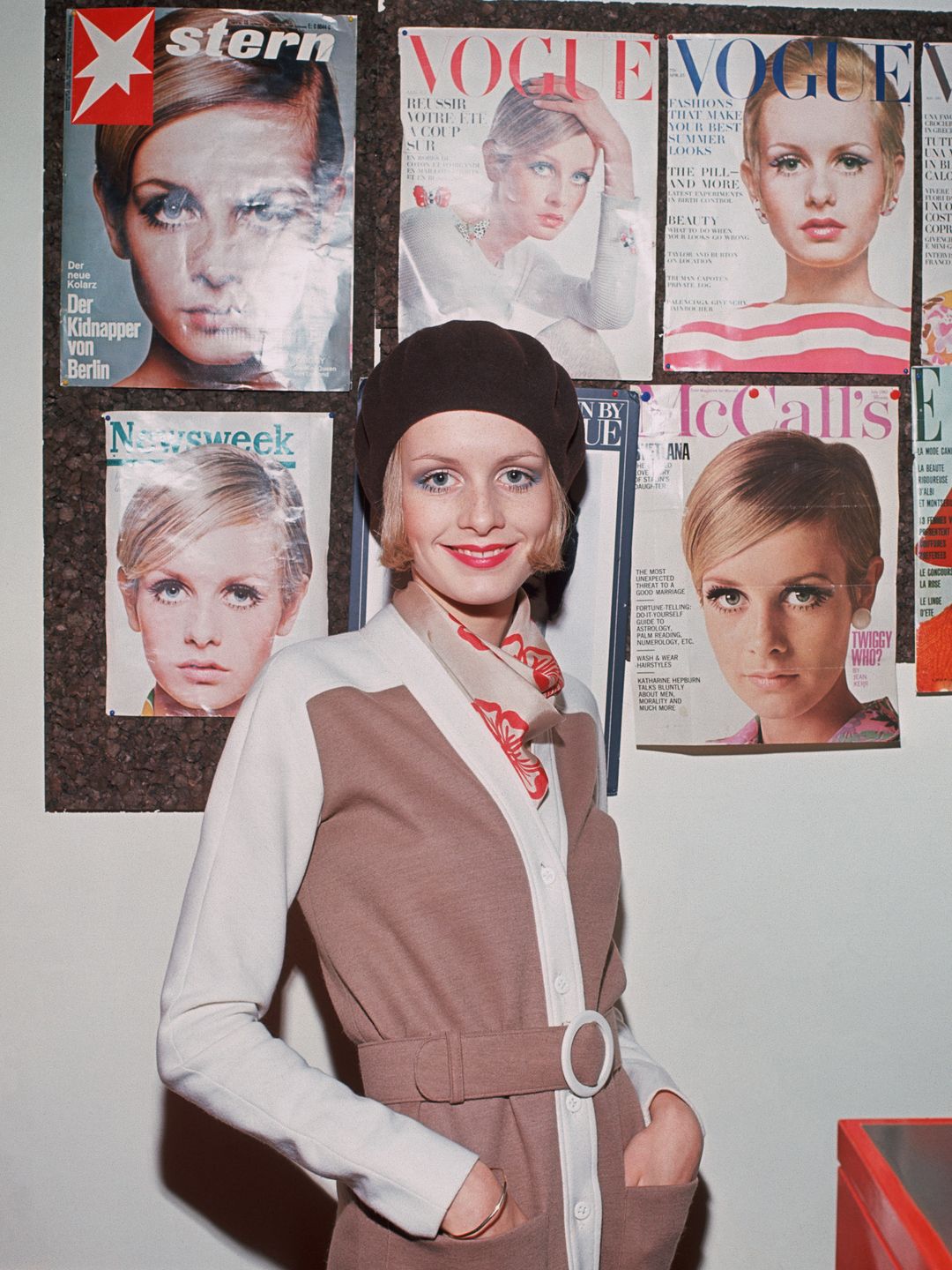 Twiggy beams beside a wall of magazine covers that celebrate her iconic status. Each cover showcases her signature doe-eyed look and bold style. Her effortless chic in a color-blocked cardigan and beret proves why she was—and remains—a fashion muse who defined 60s beauty standards worldwide.