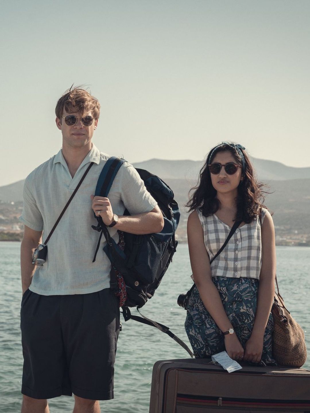 A still from Netflix's One Day Series shows actors, Leo Woodall and Ambika Mod with luggage infront of an ocean backdrop