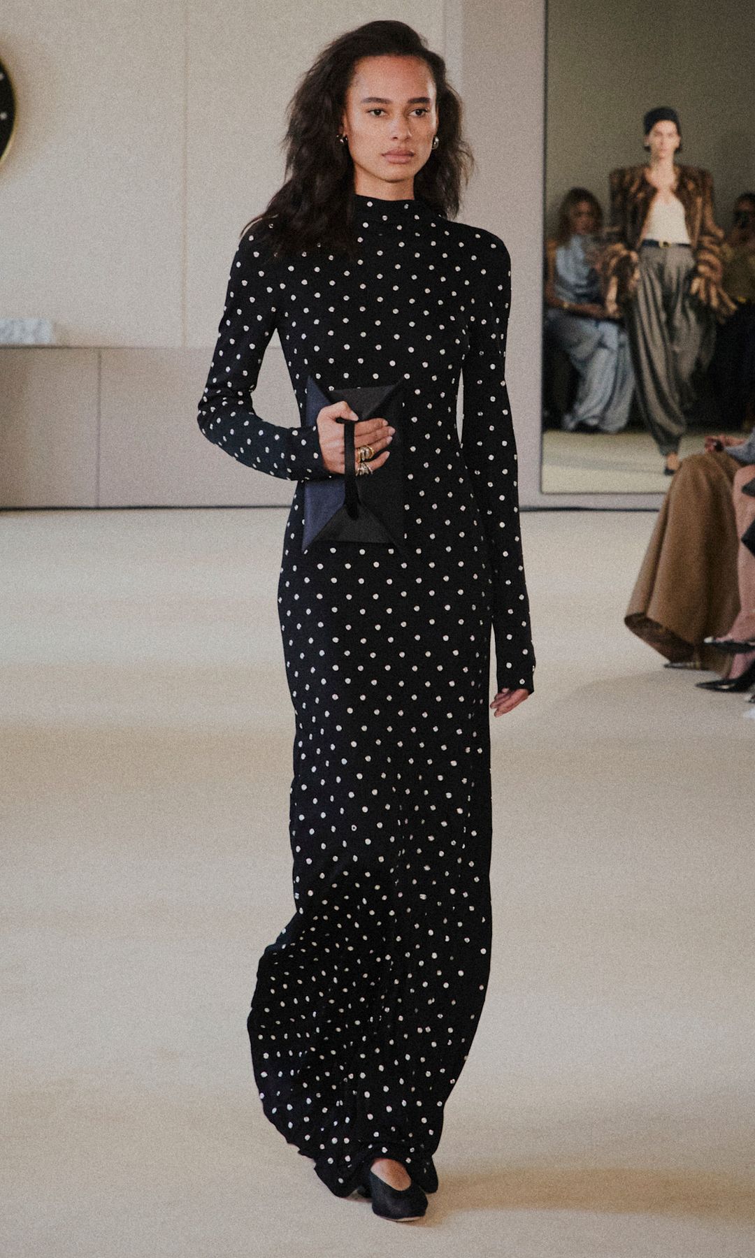 A model walks the runway in a high neck long sleeve black dress with white polka dots holding a black clutch bag
