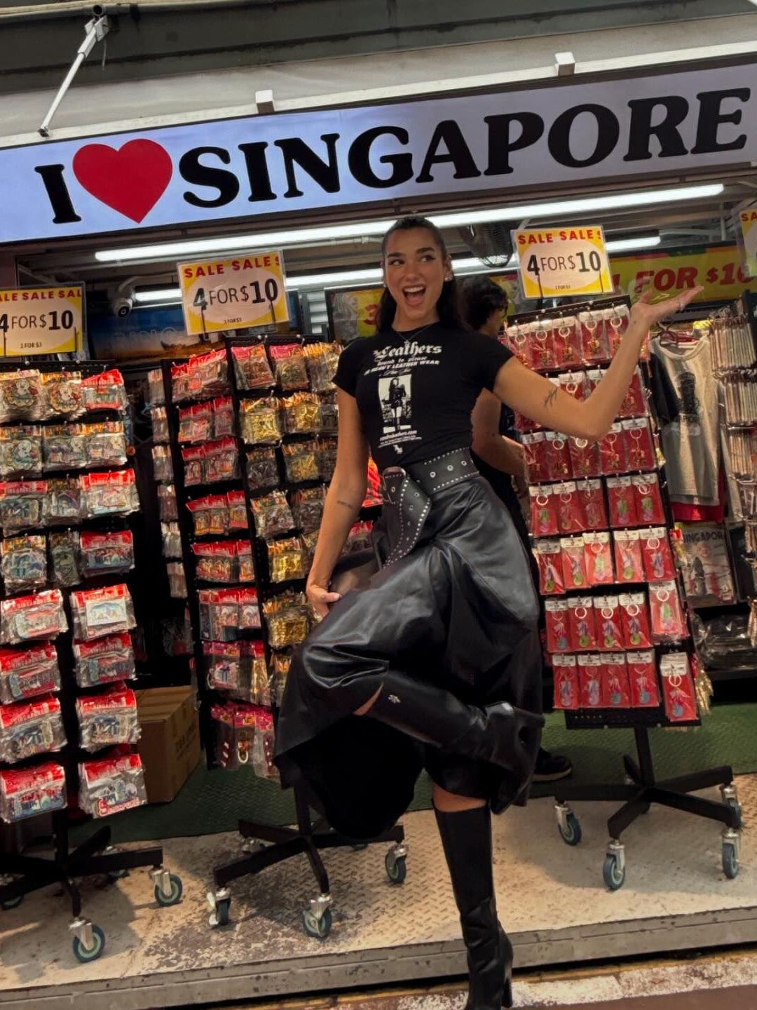 Dua Lipa poses in a black T-shirt, leather midi skirt and knee-high boots in front of a store in Singapore