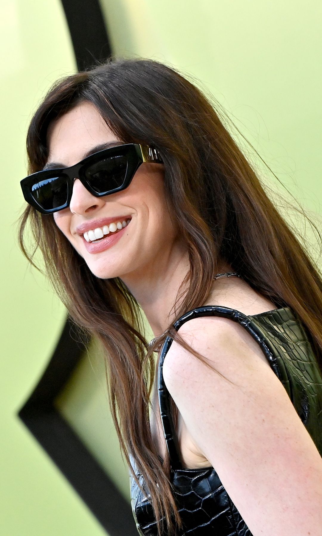 Anne Hathaway stands up against a soft Chartreuse with oversized shiny lettering and a pale lime gradient background. She wears a short, worn black mini dress with a high shiny crocodile texture. Her legs are covered in thin black tights, and she wears ultra-high black platform boots with metal buckles. A refined black handbag with a gold clasp hangs from her hand, and she finishes the look with a silver necklace, bracelet and black sunglasses. Her long brown hair is styled straight in the middle.