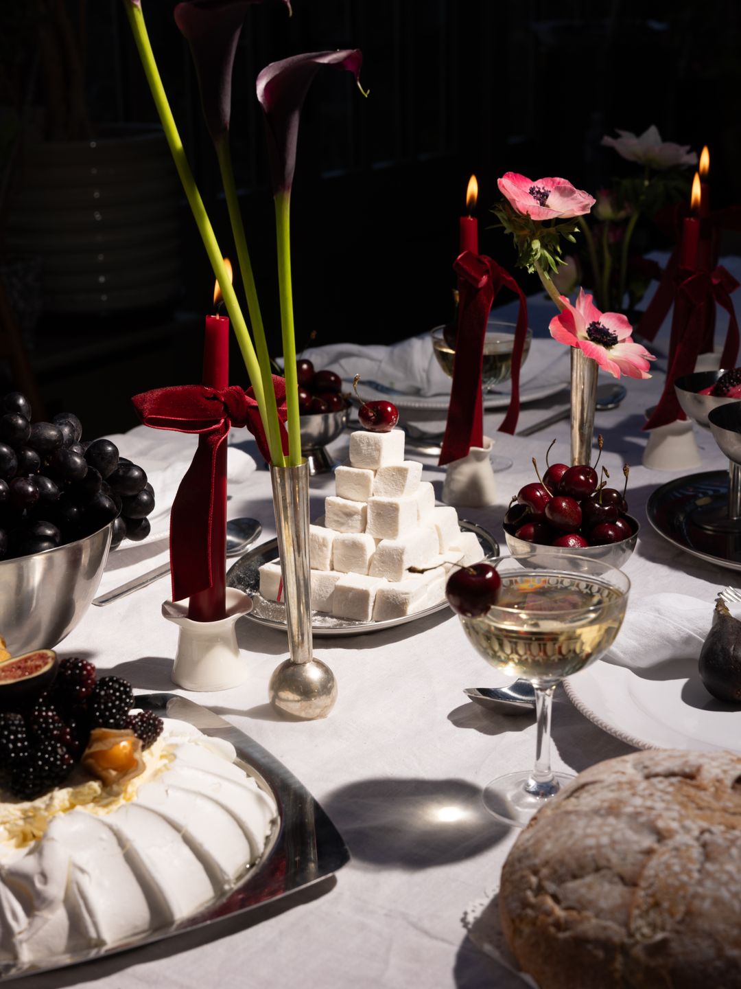 A luxurious dining table features red velvet-tied candles, dark purple calla lilies, and pink anemones in silver vases. Centered is a pyramid of marshmallows, cherries, and a cake topped with blackberries and figs. Grapes, rustic bread, and a crystal glass of wine complete this opulent, romantic feast setting.