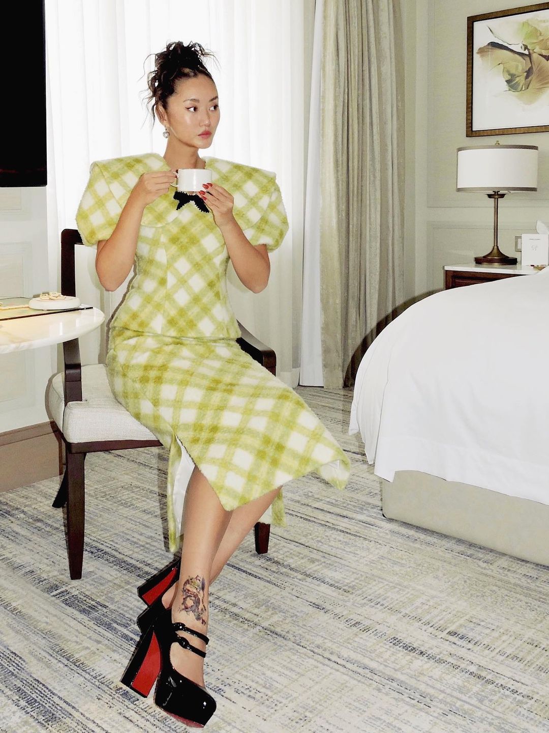 Betty Bachz sits on a chair in a hotel room sipping tea and wearing a green gingham two piece with black strapy heels