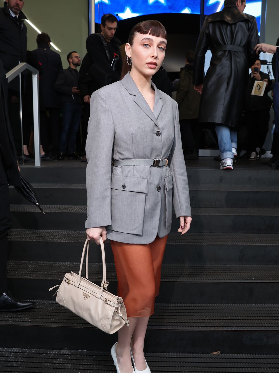 Emma Chamberlain showed up to Prada in a grey belted blazer, orange sheer shirk, white heels and a micro fringe. 