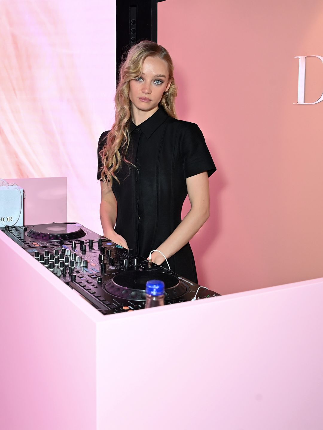 DJ Maddi Waterhouse in a sleek black outfit spins tracks at a pink Dior-branded booth. Behind her, a soft-lit display showcasing beauty imagery creates a modern, immersive atmosphere at this high-profile brand event.