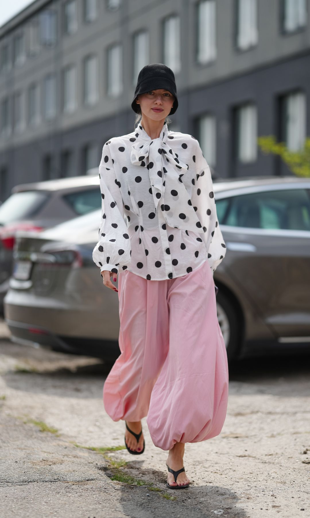 A guest wears a black bucket hat with light blonde hair tucked underneath, soft pink lipstick, a white lightweight woven blouse with oversized black polka dots, a self-tie bow at the neck, and billowy long sleeves with gathered cuffs, paired with voluminous pale pink balloon-hem trousers in a flowy fabric gathered at the ankles, black flat flip-flops with thin straps, and bright red nail polish on fingernails, walking, outside Marimekko, during Copenhagen Fashion Week,