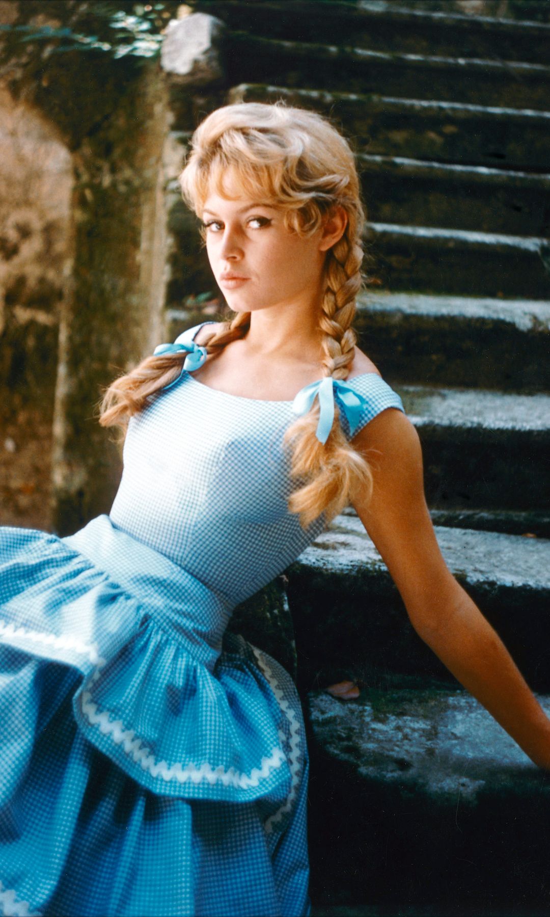 Brigitte Bardot leans against some steps wearing a blue gingham dress and pigtails tied with blue bows.