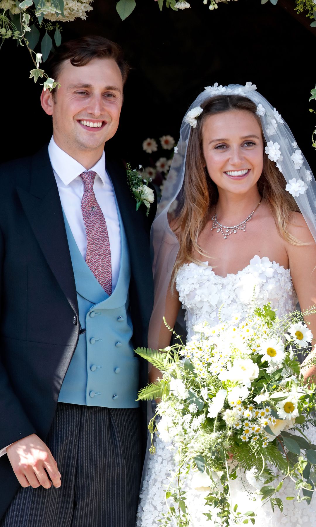 The bride and groom. Charlie van Straubenzee and Daisy Jenks. Daisy wears a long embroidered veil with a strapless wedding dress and holds a rustic bouquet of daisies