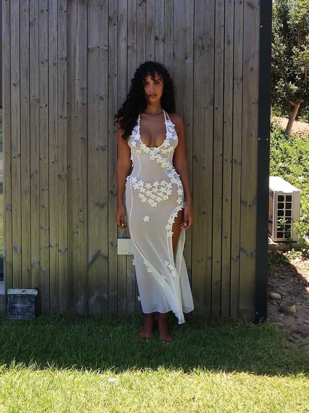 Maya Jama Poses infront of a wood house wearing a sheer white dress