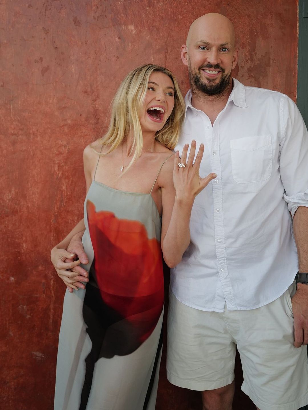 Georgia Toffolo joyfully displays her engagement ring, laughing beside James. She wears a dress with a large red floral design, and they stand against a reddish wall, looking delighted about their engagement.