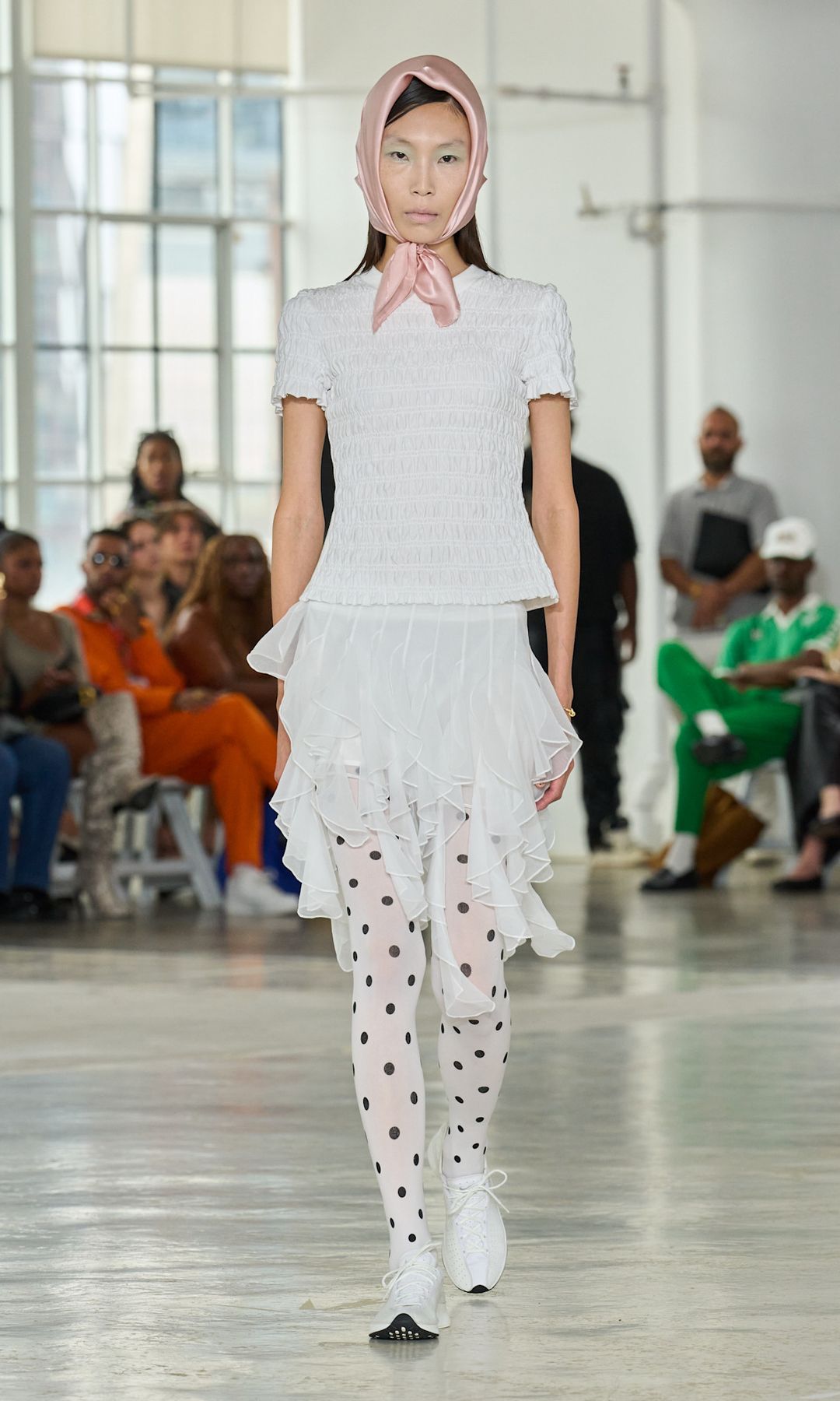 A model wears a white shirred top, white ruffled mini skirt, polka dot tights and pale pink headscarf