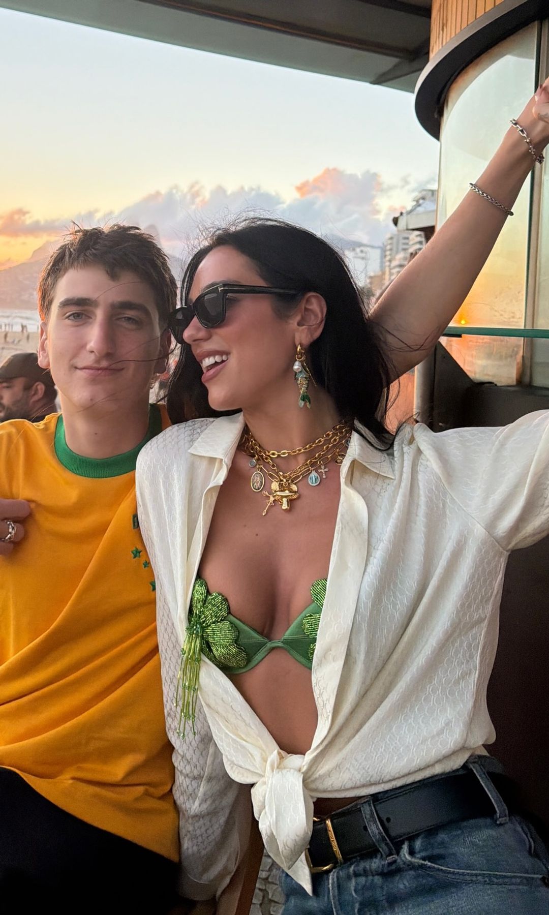 Dua Lipa poses with her brother on a balcony wearing jeans, a white shirt and green beaded bra 