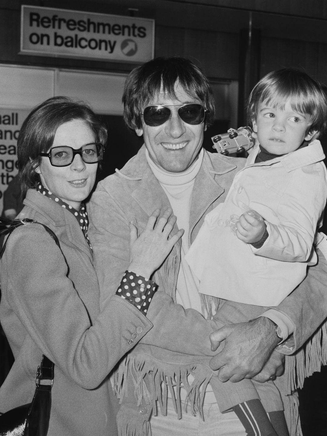 This photo shows Maggie Smith with her family in a casual setting. She wears glasses and a stylish 1970s outfit while holding her young child. Her partner stands beside her, sporting sunglasses and a fringe jacket. Maggie’s expression is calm, and the overall vibe is one of effortless family cool. It’s a candid, intimate moment that reflects the laid-back, stylish aesthetic of the time.