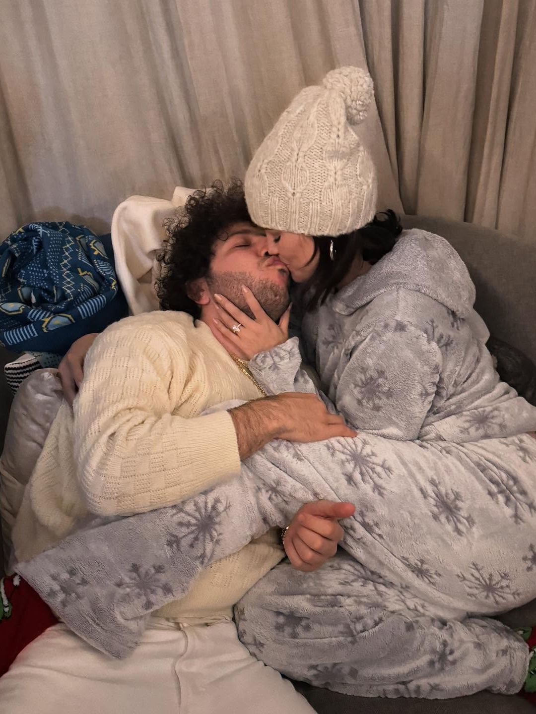 Selena Gomez and Benny Blanco sit cuddled up on a grey armchair together, sharing a kiss. Selena is wearing a grey tracksuit with snowflakes on it and a cream hat. Benny is wearing a cream jumper and white trousers. The image is romantic, warm and cosy in nature. 