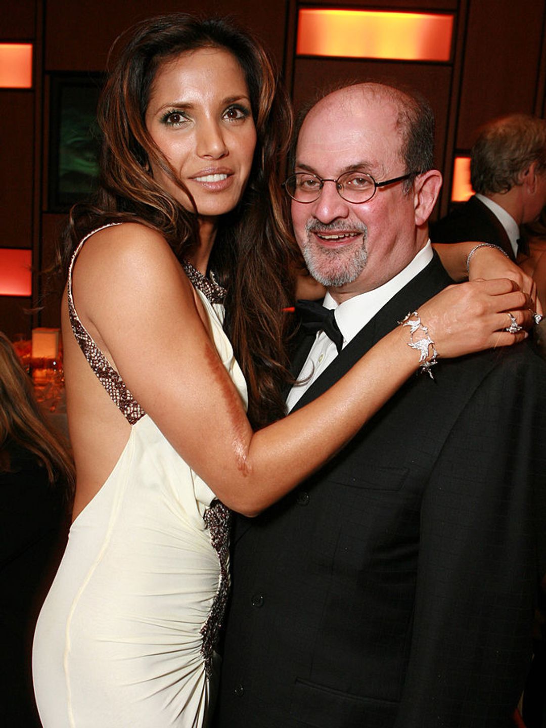 Padma Lakshmi and Salman Rushdie at a party. She is wearing a white backless dress and he is dressed in a classic tuxedo. 