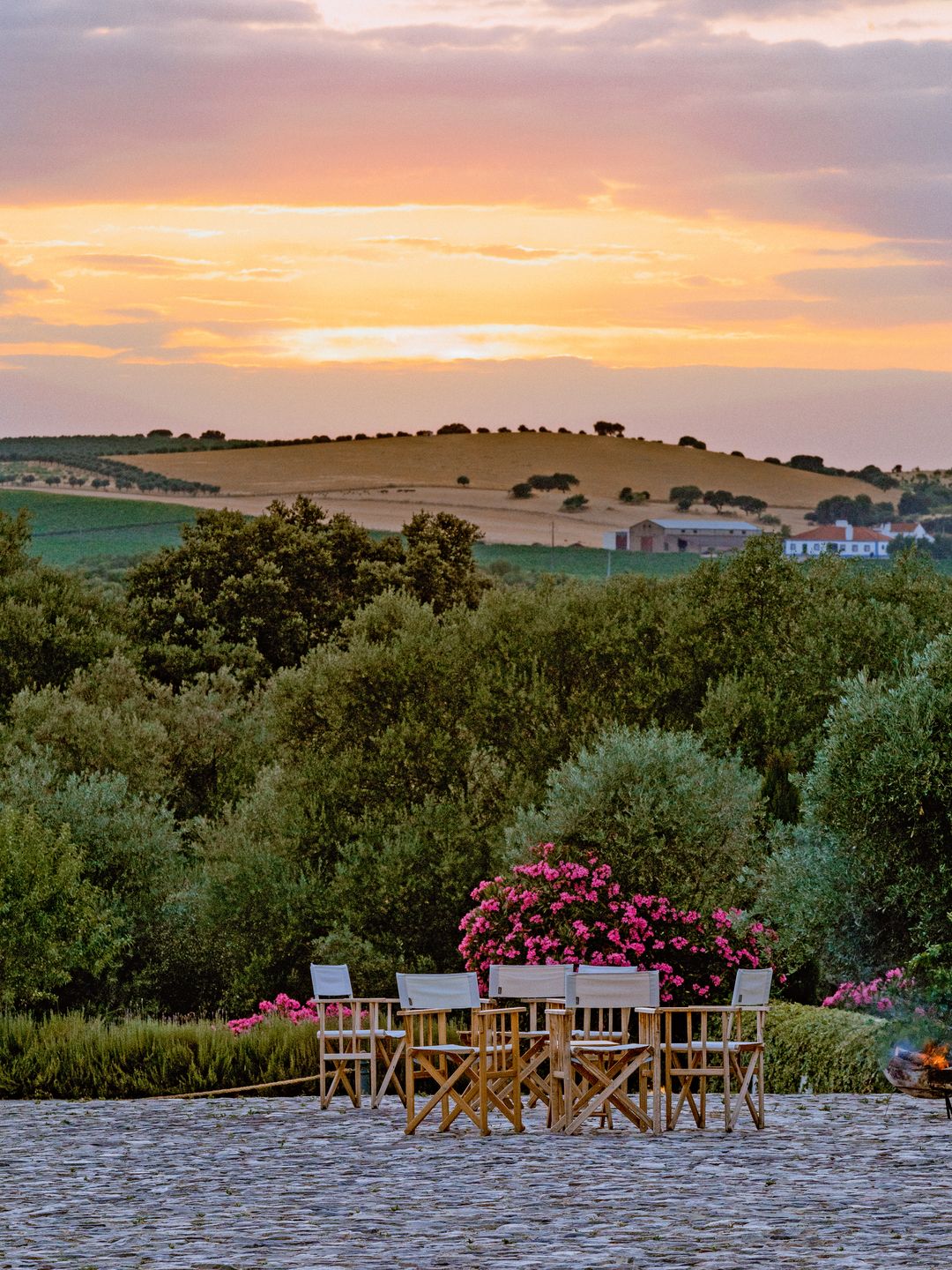 A picturesque sunset over rolling hills and green trees, with a cobblestone patio in the foreground featuring a cluster of white wooden chairs around a table.