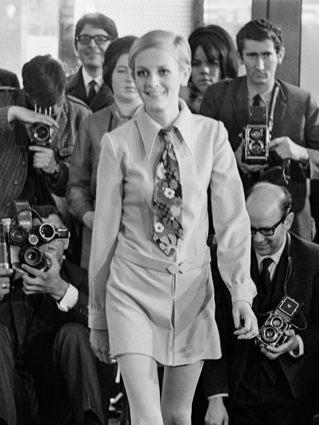   Twiggy ma na sobie szykowną mini-sukienkę i kwiatowy krawat. Skupienie się fotografów na niej odzwierciedla jej status ikony mody lat 60., znanej z redefiniowania standardów piękna i wyznaczania trendów swoim nowoczesnym, androgynicznym wyglądem.