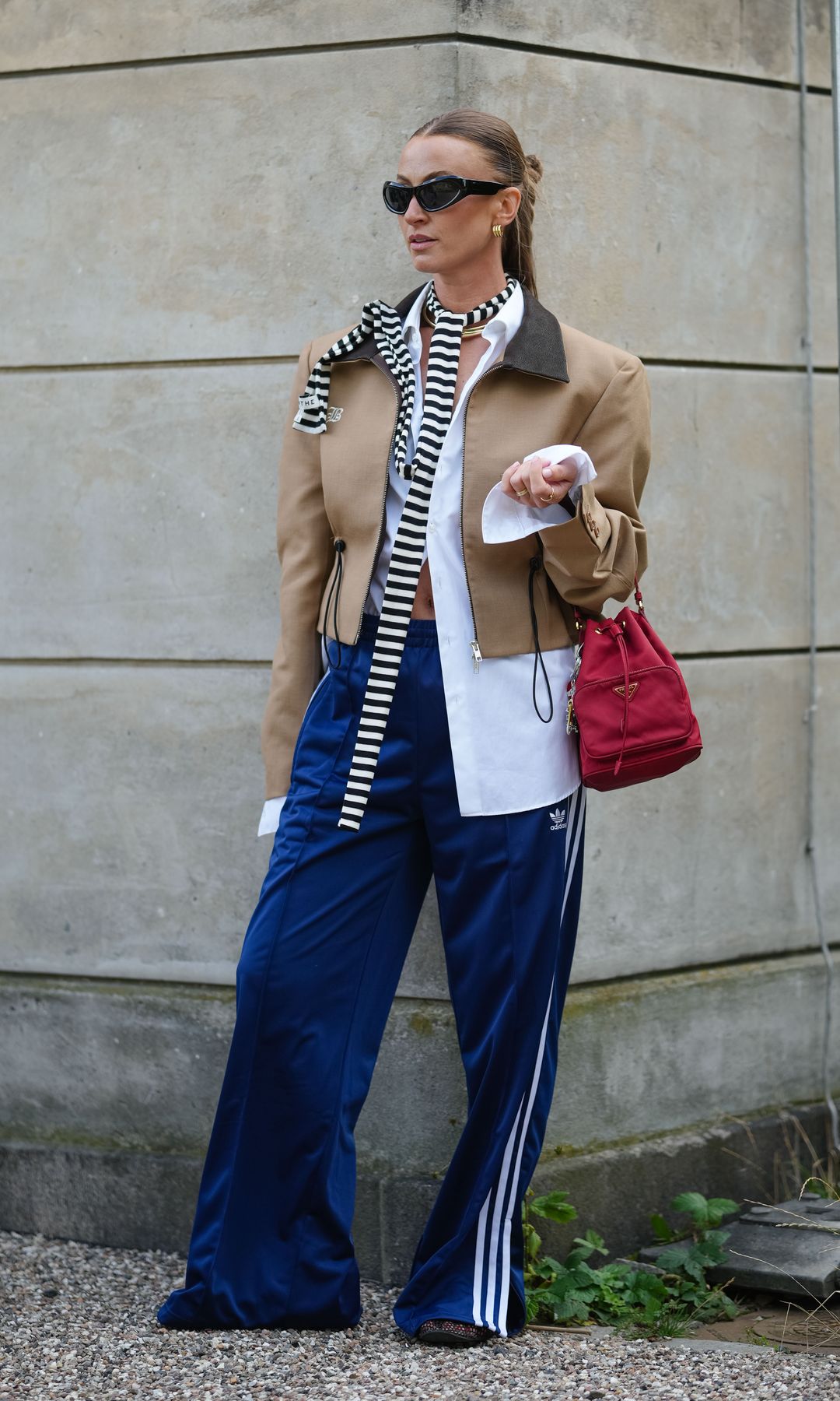 A guest wears a camel cropped jacket with a dark brown collar and zip fastening, layered over a white shirt with exposed cuffs. A black and white striped scarf is draped around the neck. Blue Adidas track pants with white side stripes and a visible logo are worn. Accessories include black cat-eye sunglasses, gold earrings, and a red handbag with a triangular logo and gold hardware. Hair is styled in a sleek, pulled-back braid, outside Munthe, during Copenhagen Fashion Week