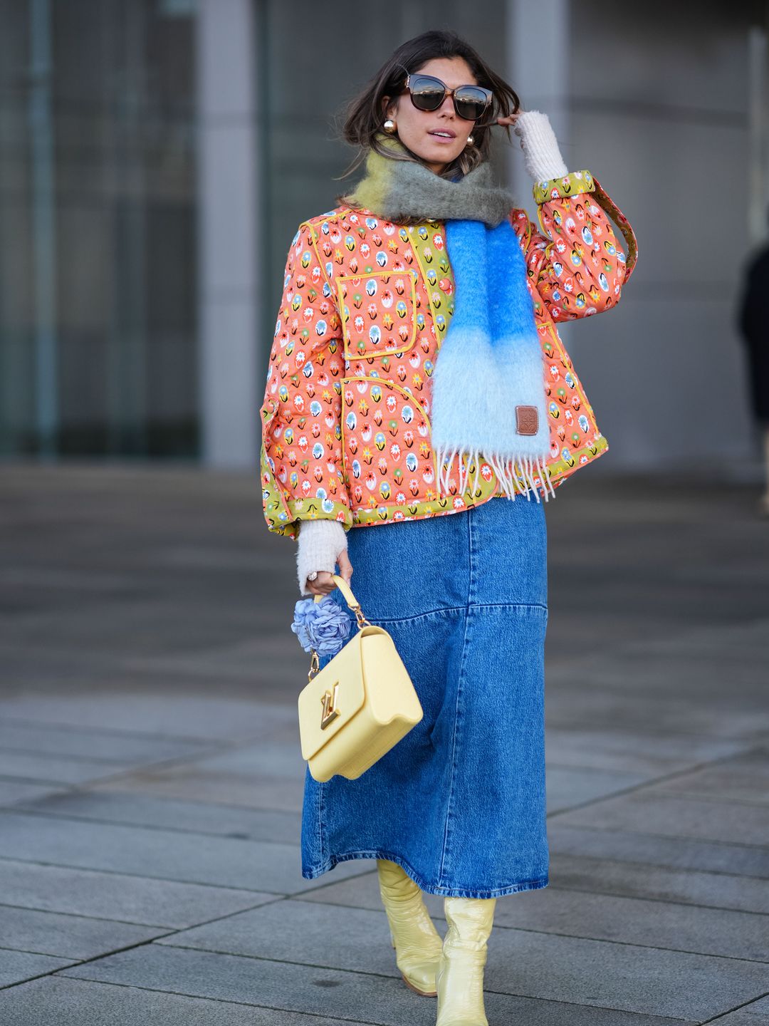 A guest wears sunglasses, a khaki / gray / blue wool fringed scarf, an orange floral print padded jacket with yellow lines, a pastel pale Vuitton bag, a blue denim midi skirt, high heels leather boots , outside OperaSport, during the Copenhagen Fashion Week AW24