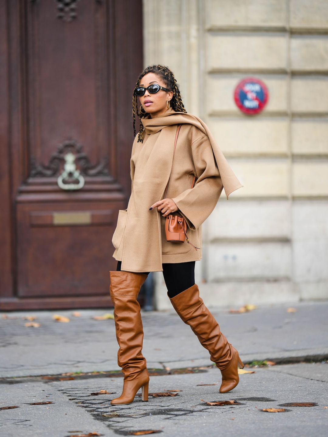 Ellie Delphine wears a camel coat with black leggings and tan thigh-high boots 