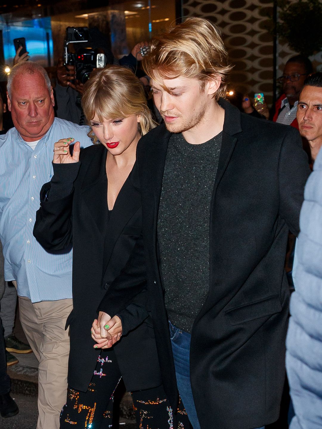 Taylor and Joe walking through a crowd with security around them, keeping their heads angled down, they are holding hands