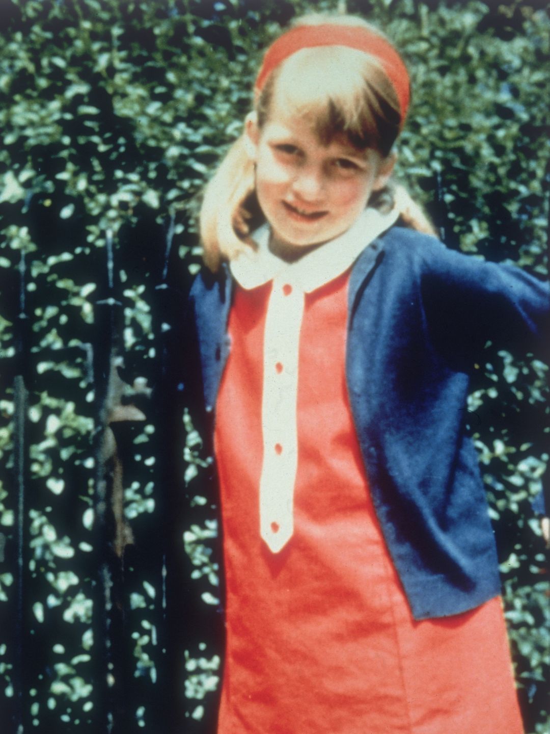 A young Princess Diana in a red dress and hairband with blue cardigan