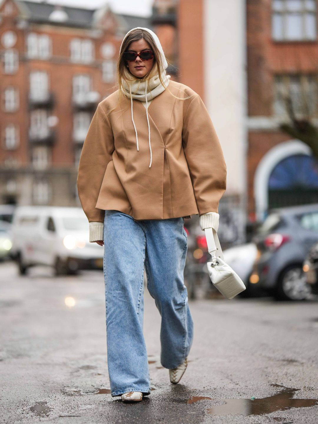 Woman wearing beige ribbed wool hoodie, a beige wool pullover, a brown oversized jacket, blue faded denim large pants, a white shiny leather handbag