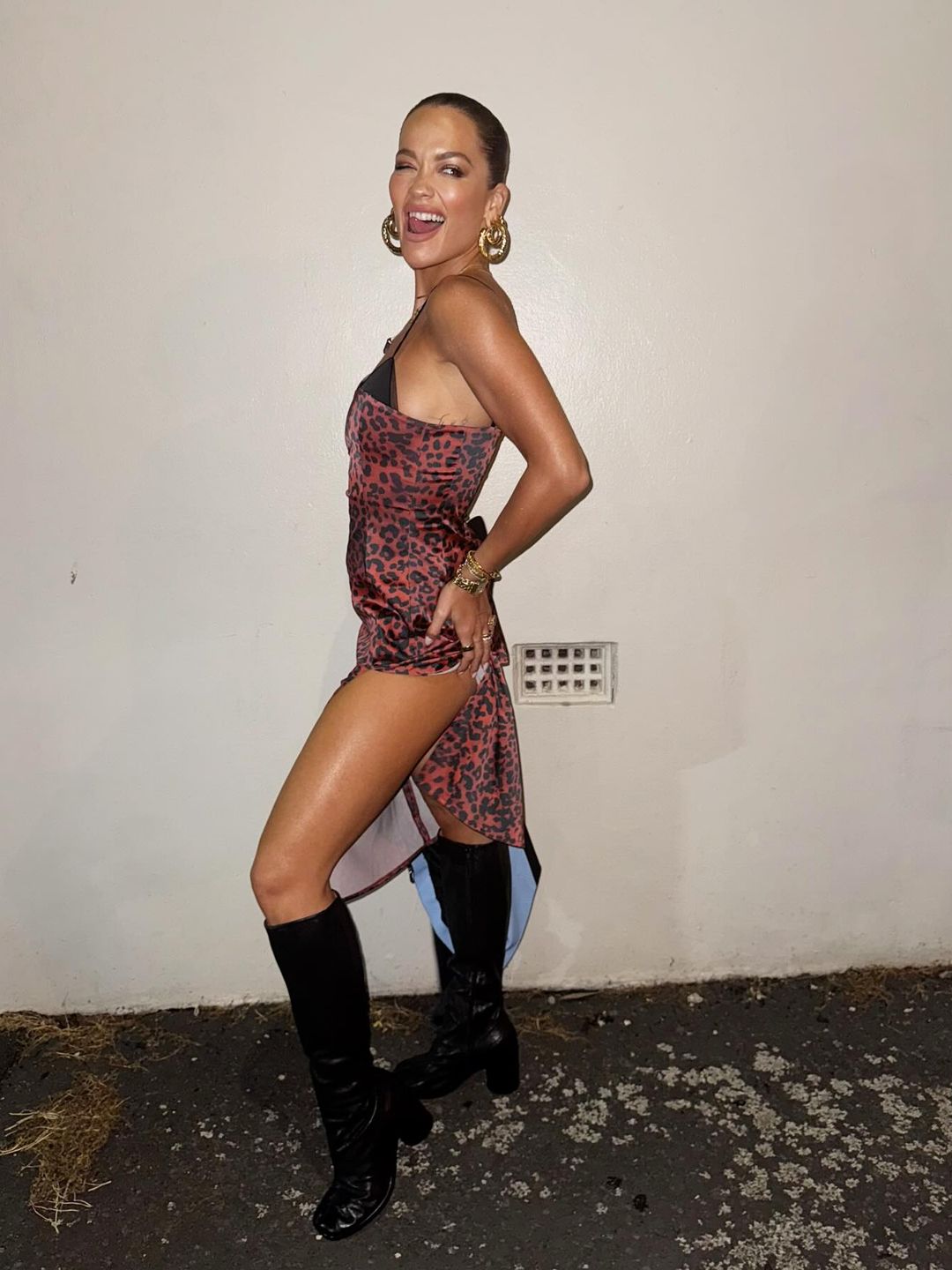 Rita Ora wears a dark red leopard print slip dress with big gold earrings and black knee high leather boots. She winks playfully at the camera. 