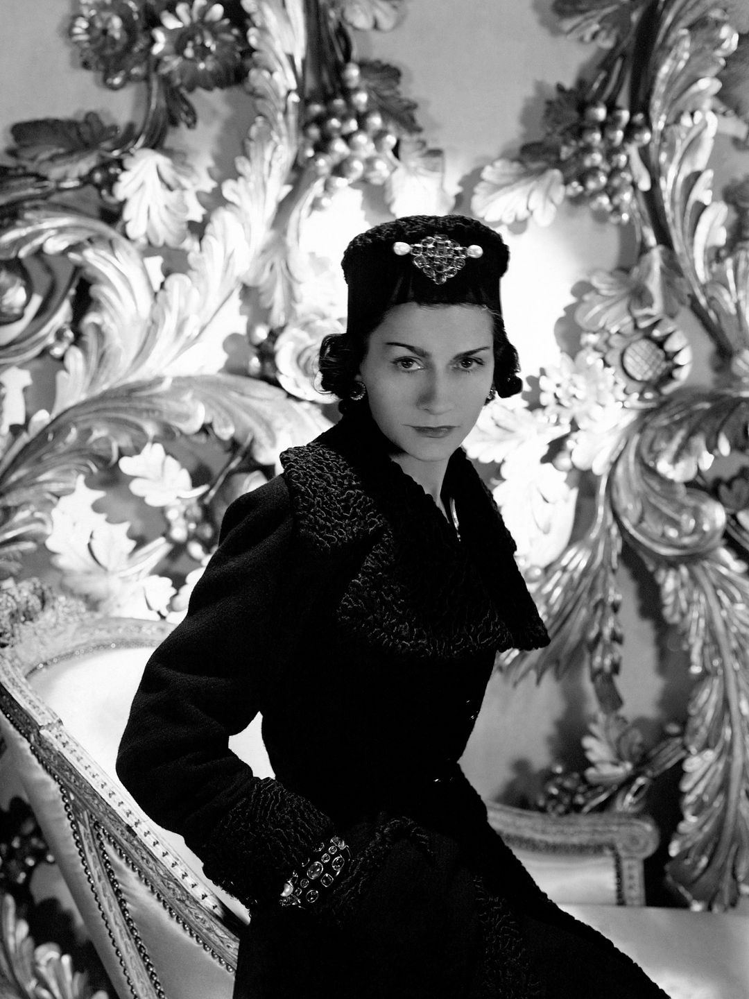 Coco Chanel wearing one of her designs: a three-quarter coat with cinched waist and astrakhan lapels and cuffs, and matching astrakhan hat with large brooch on front. 