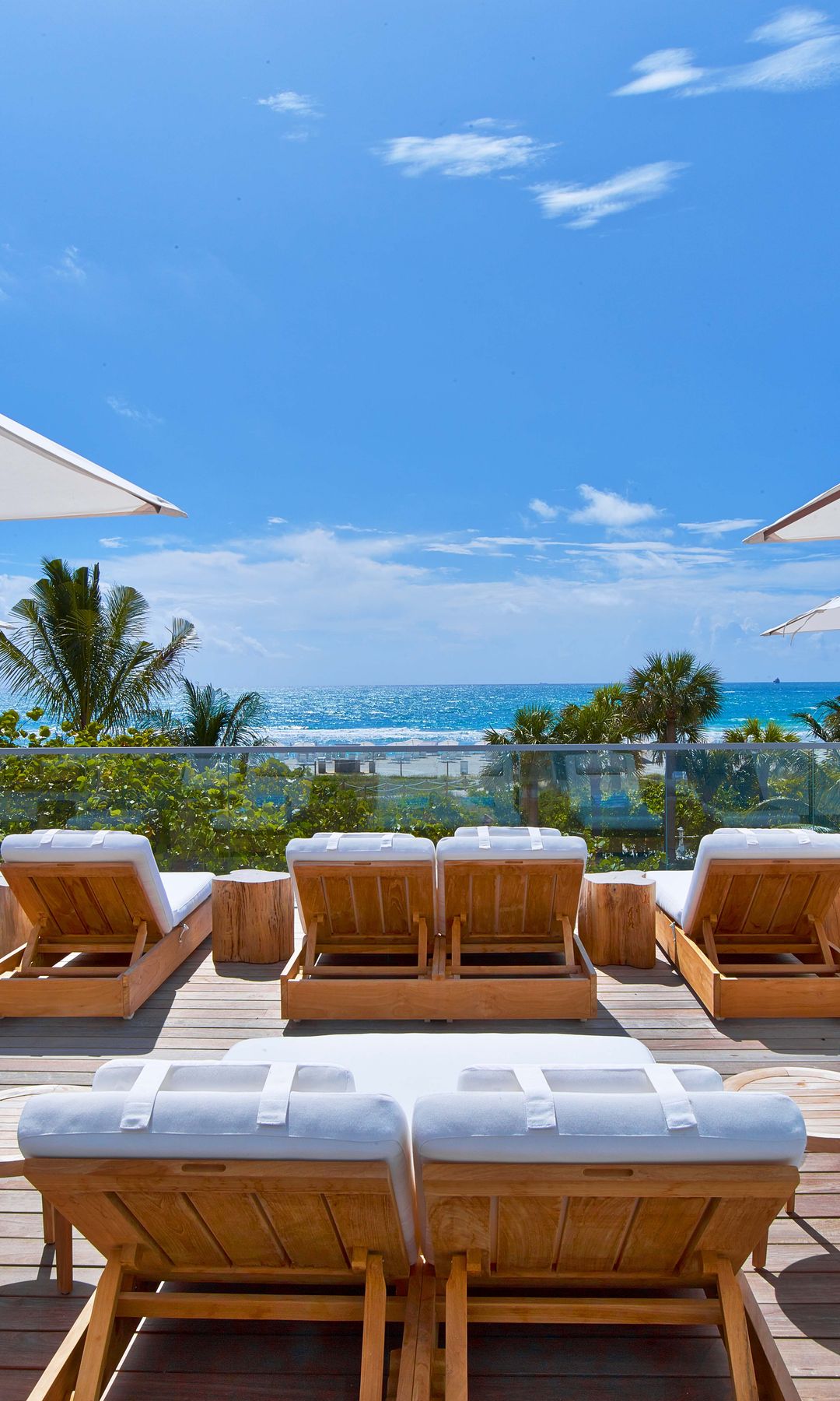 A bright, sunlit rooftop deck overlooking the ocean, featuring rows of wooden sun loungers with plush white cushions.