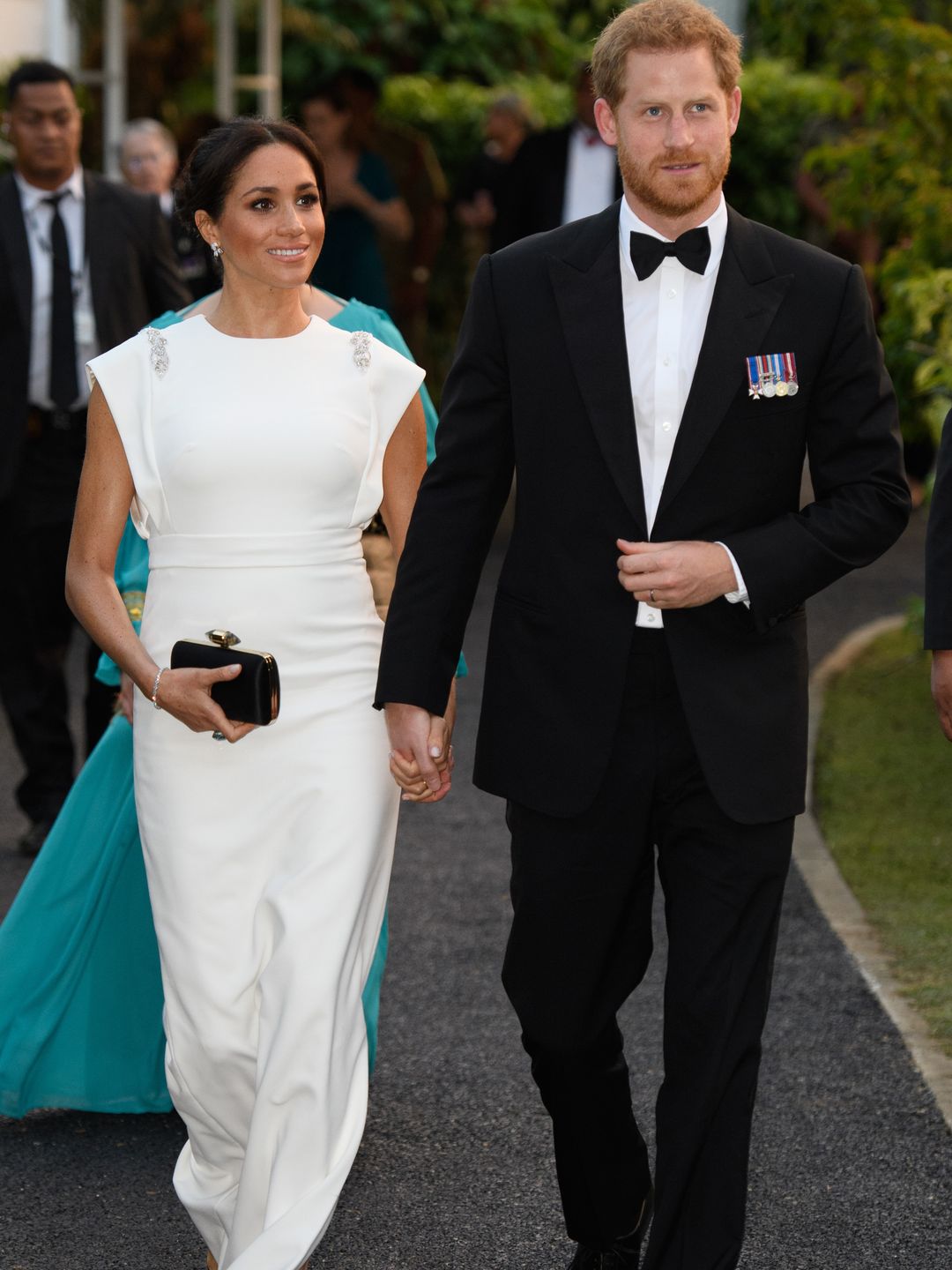The Duchess of Sussex in a white dress holding hands with Prince Harry