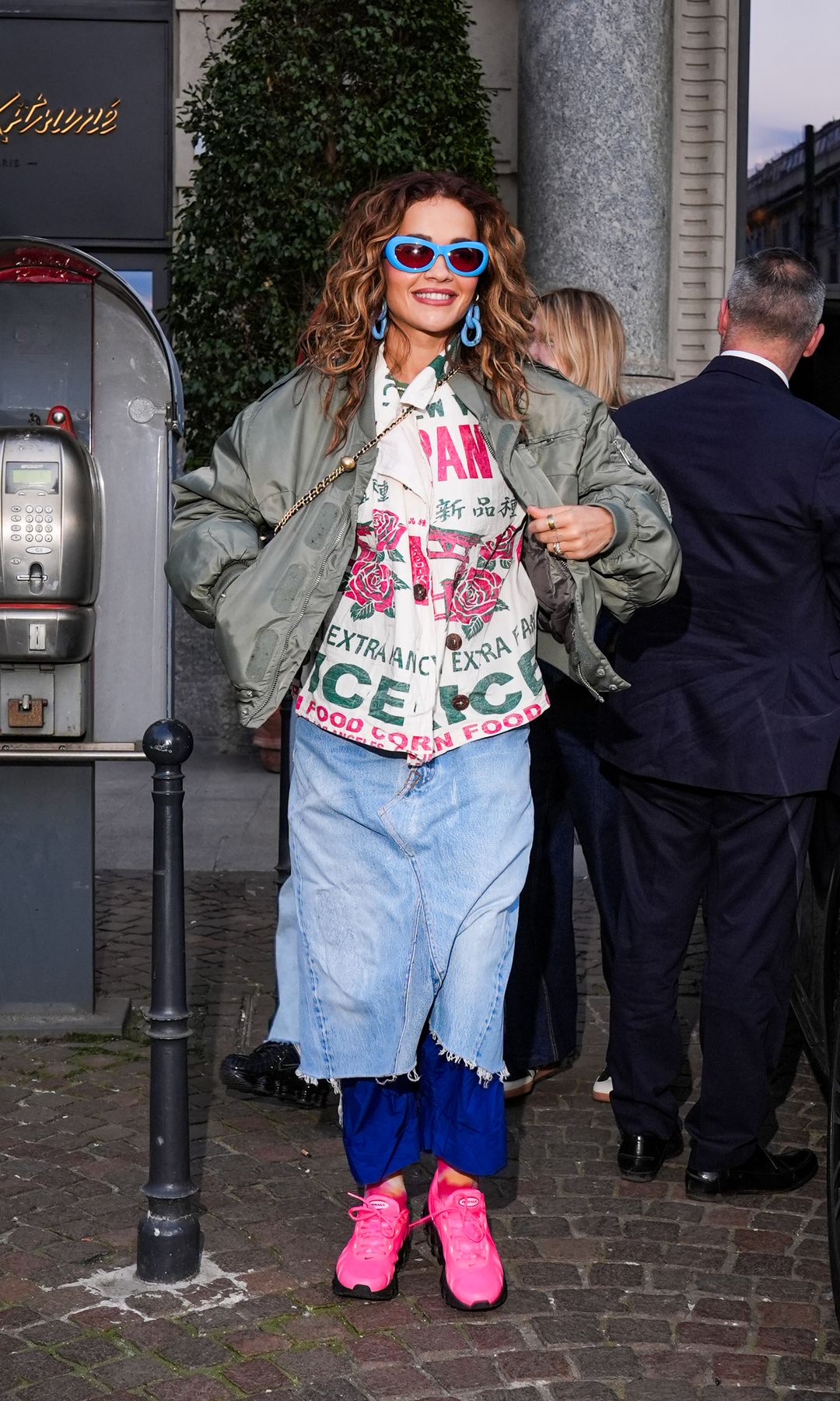 Rita Ora is seen wearing a denim skirt over blue torusers, a button-up graphic shirt and green bomber jacket while outside her hotel in Milan, Italy