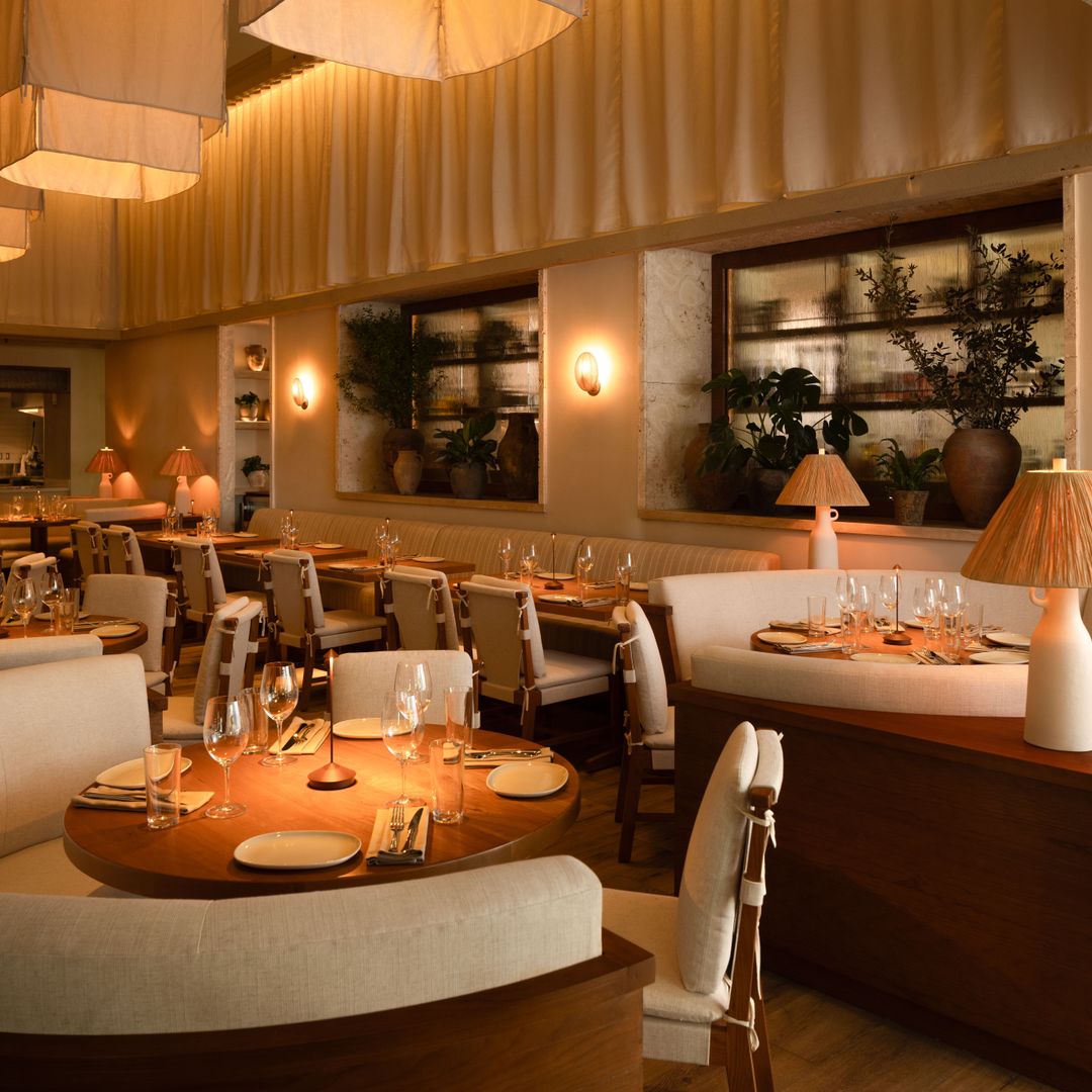 The interior of AVIV, a refined Mediterranean-inspired restaurant at 1 Hotel South Beach. The space features curved cream banquettes and wooden dining tables set with elegant glassware and neutral-toned place settings.
