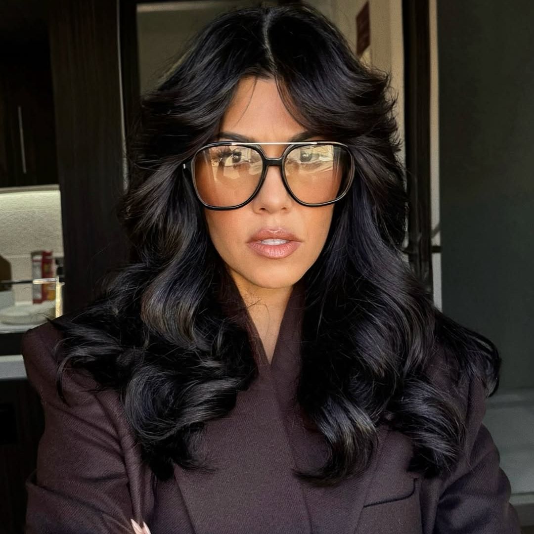Kourtney Kardashian with blakc 70s hair and a brown blazer 