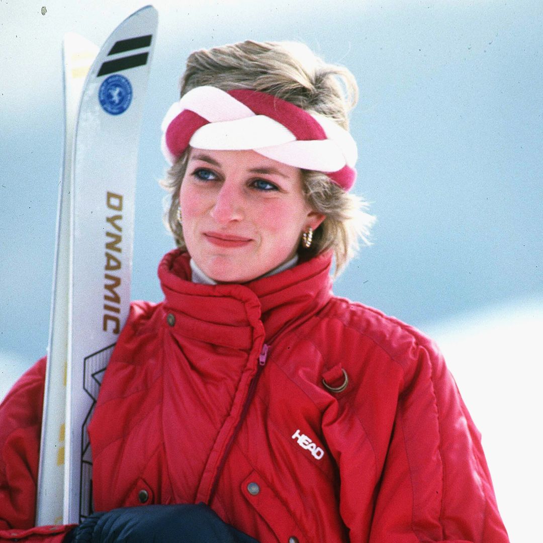 The image depicts a woman outdoors in a snowy setting, wearing a bright red ski jacket with "HEAD" visible marking on the chest. She is holding a pair of skis and ski poles, and wearing black insulated gloves to keep warm. Her blonde hair is short and she wears a red and white braided headband on her head. Her expression is calm and slightly smiling, and she looks ready to ski or do some cold-weather activity. The bright snow in the background enhances the wintry atmosphere of the scene.