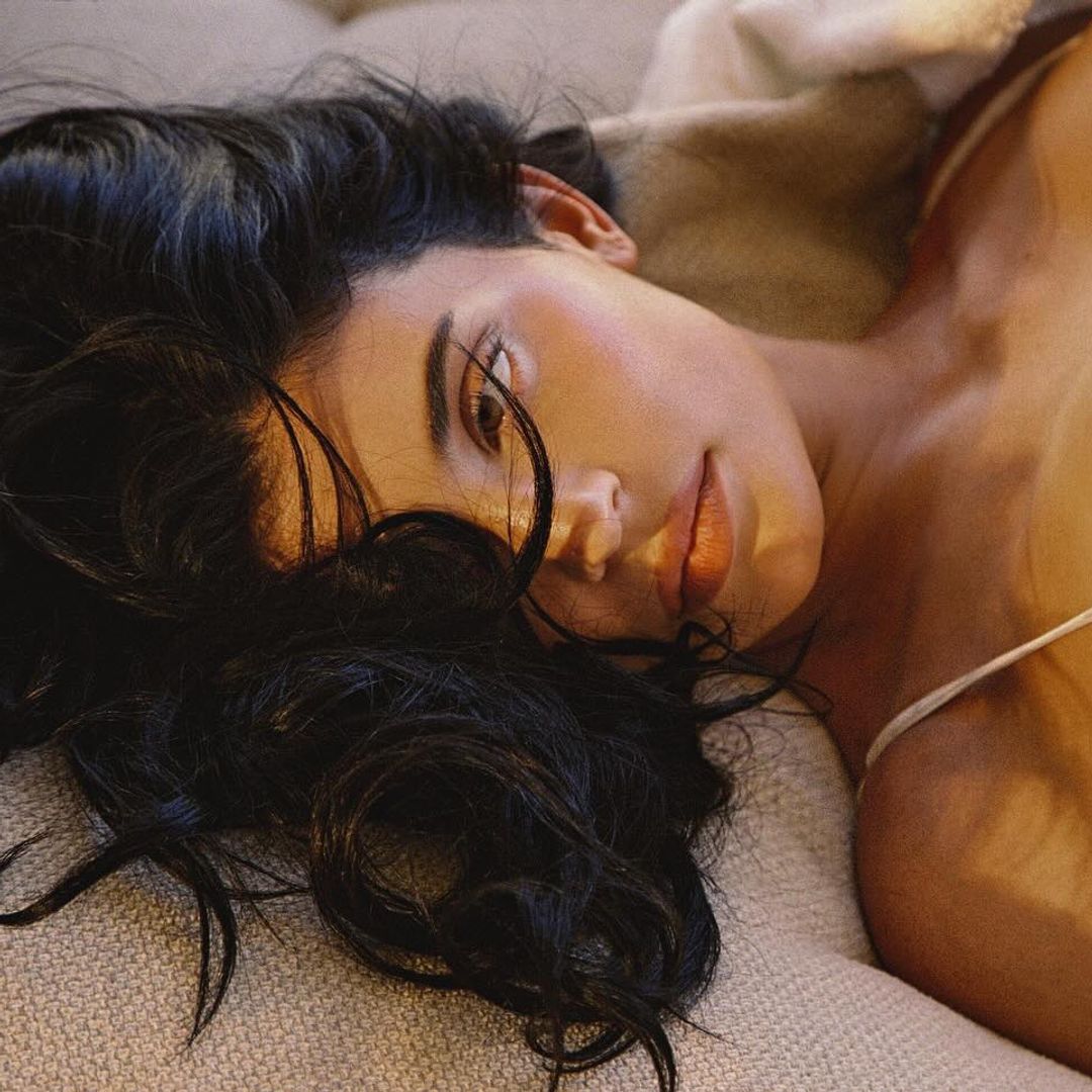 Kylie Jenner is lying on her back on a plush beige sofa, gazing directly into the camera with a serene, sultry expression. She wears a delicate white bikini that mimics vintage lingerie, featuring subtle polka-dot texture and underwire detailing reminiscent of retro brassieres. Her tousled dark hair cascades across the cushions, and her hands gently touch her chest and shoulder. Soft lighting creates a golden, intimate atmosphere, giving the scene a cinematic warmth and softness.
