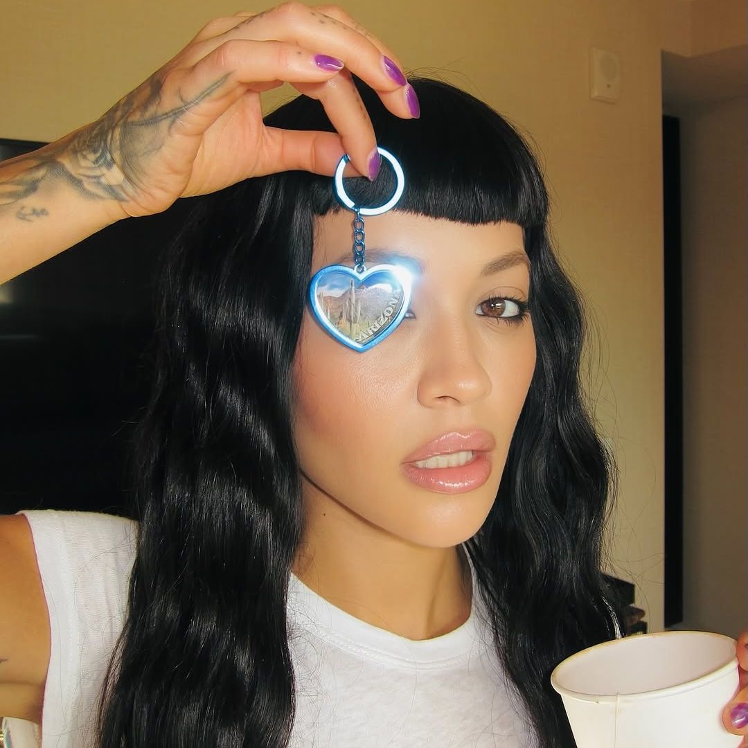 Rita Ora with a jet black micro fringe and baby bangs holding up a heart pendant 