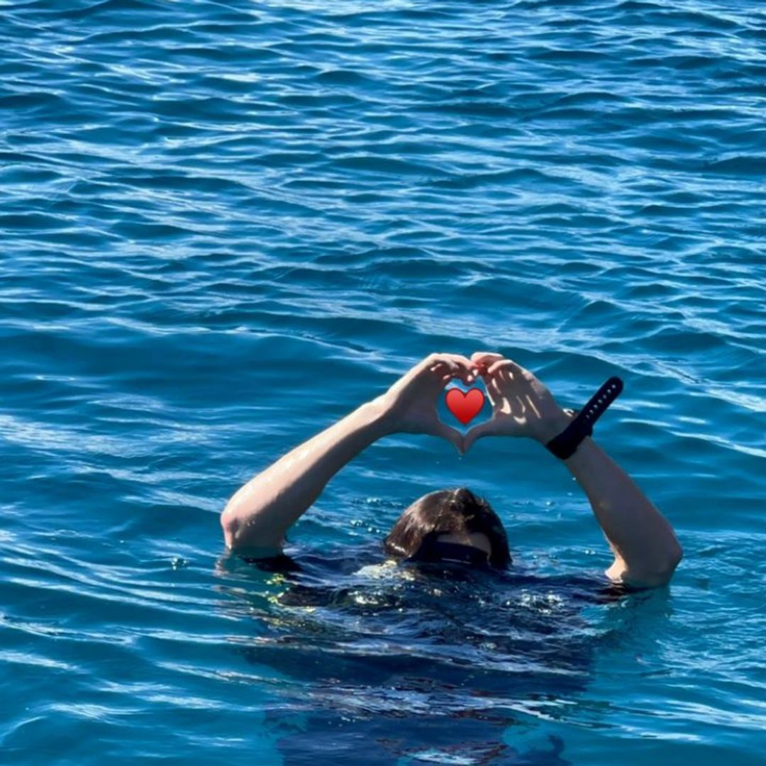 Tom Holland standing in the sea, his head is underwater but he is holding his arms out to make a heart sign