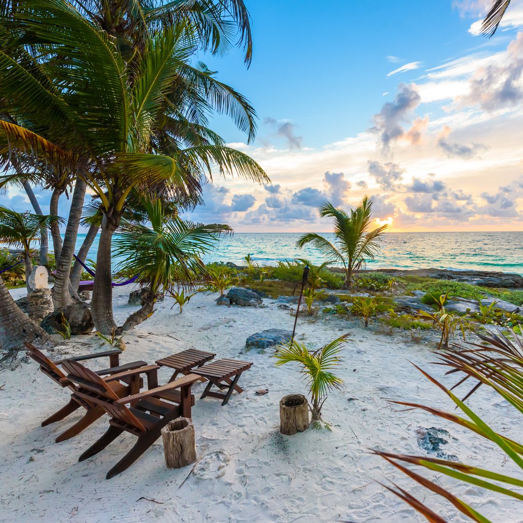 a picture of a white sandy beach, blue ocean, palm trees and two deck chairs on the sand