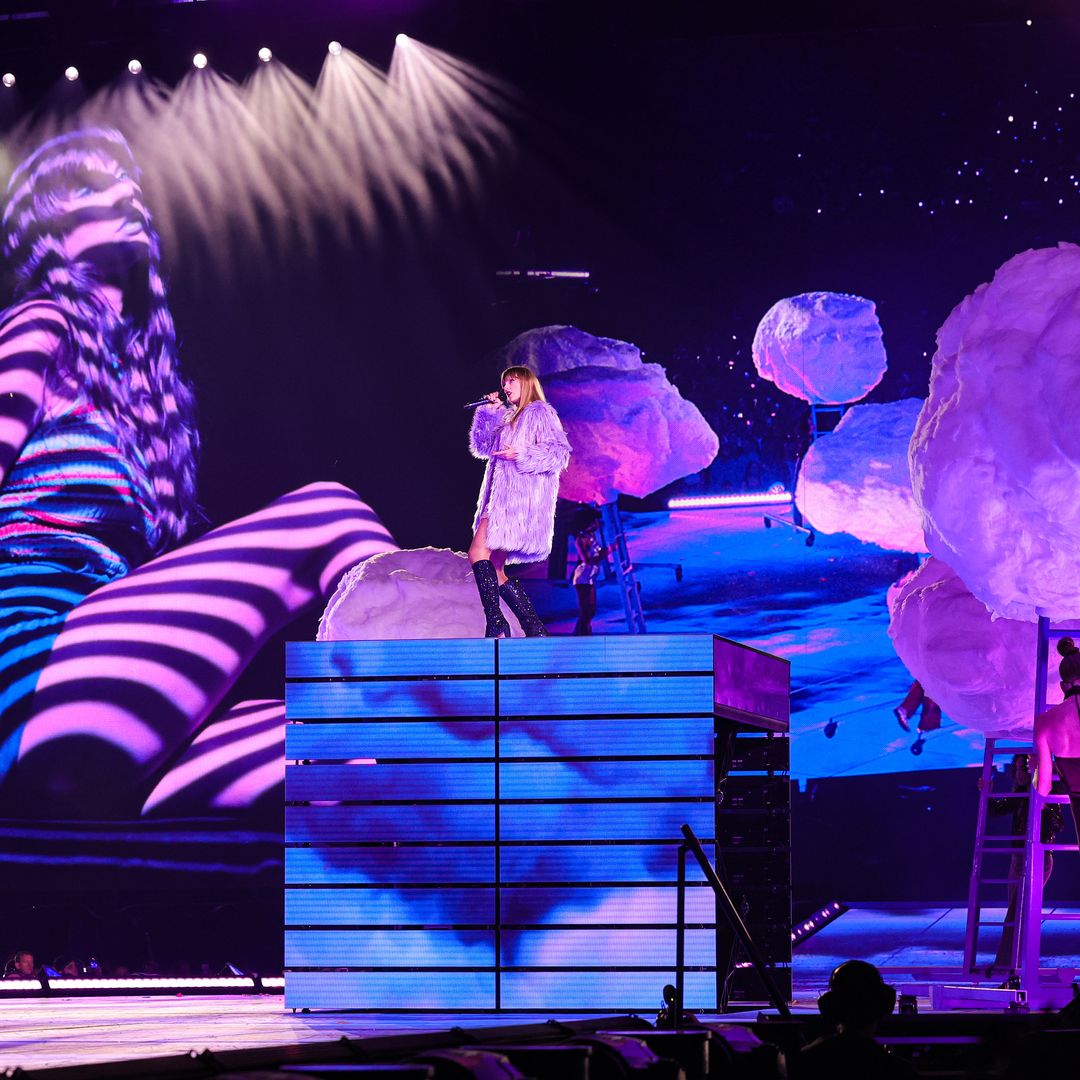 Taylor stood on a high platform singing on her tour, she is wearing a lavender colored coat and is surrounded by purple clouds and lighting