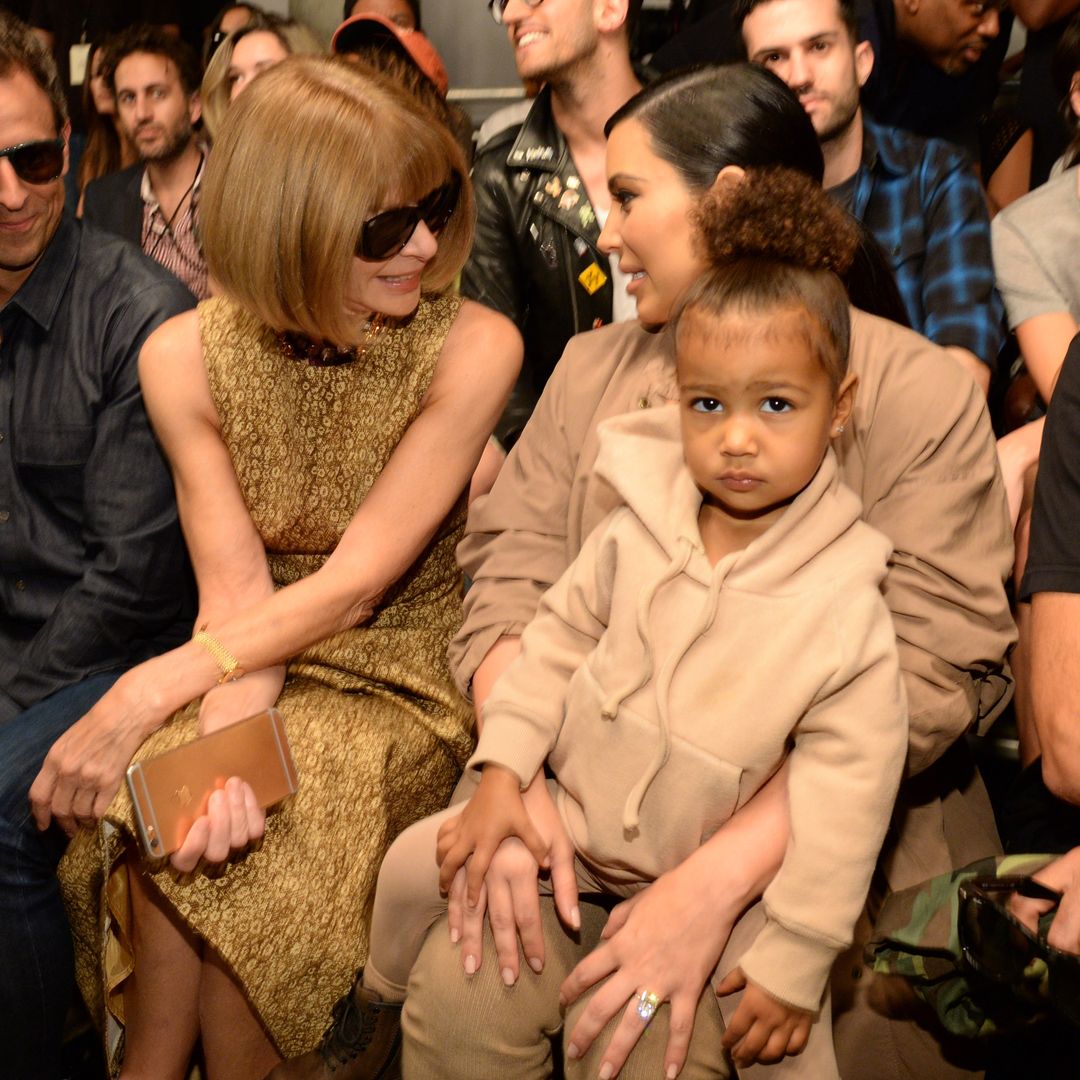 North West sat on Kim's lap at a fashion show, Anna Wintour is chatting to Kim as North looks at the camera