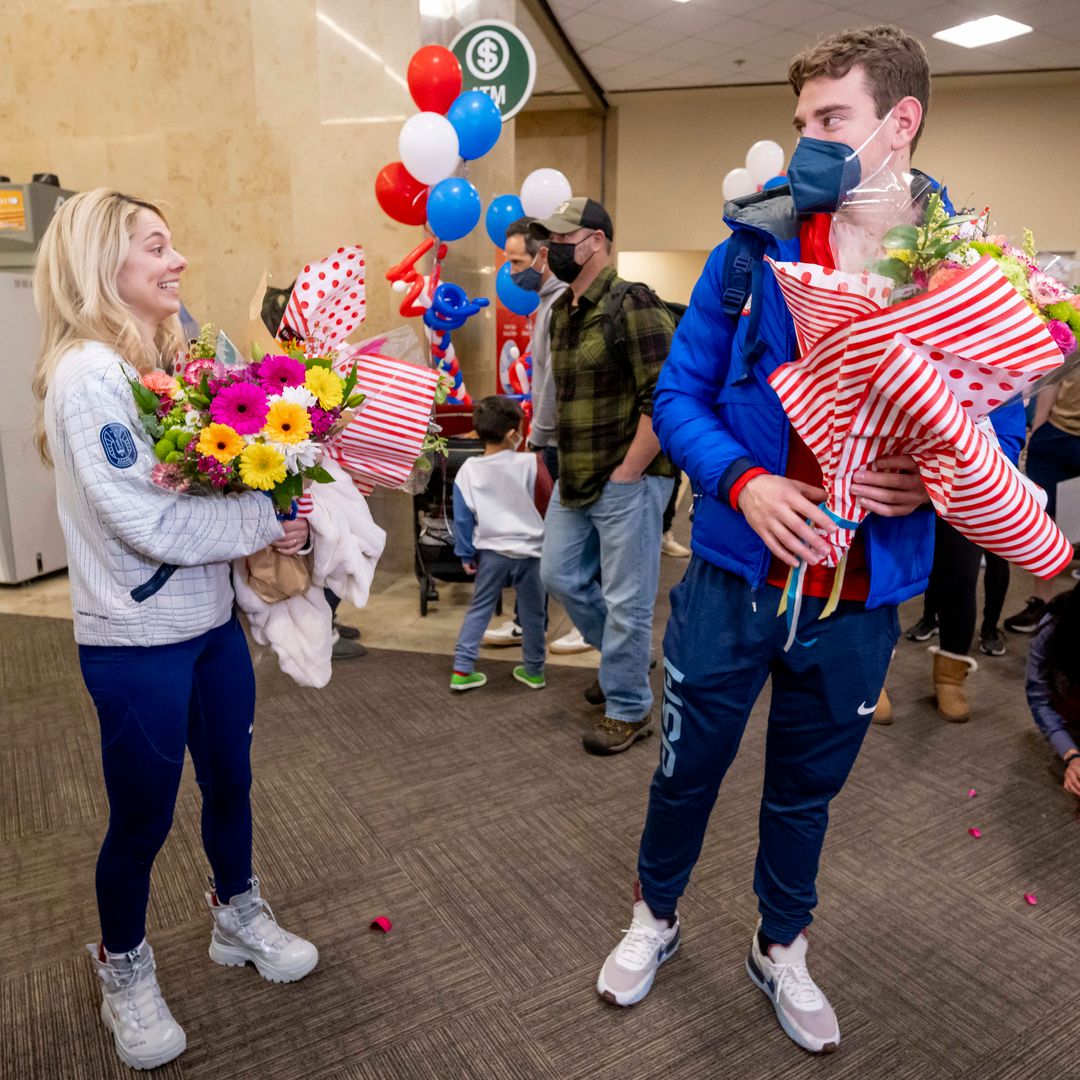 Skaters Alexa Knierim and Brandon Frazier holding flowers and flags and smiling in an airport lobby