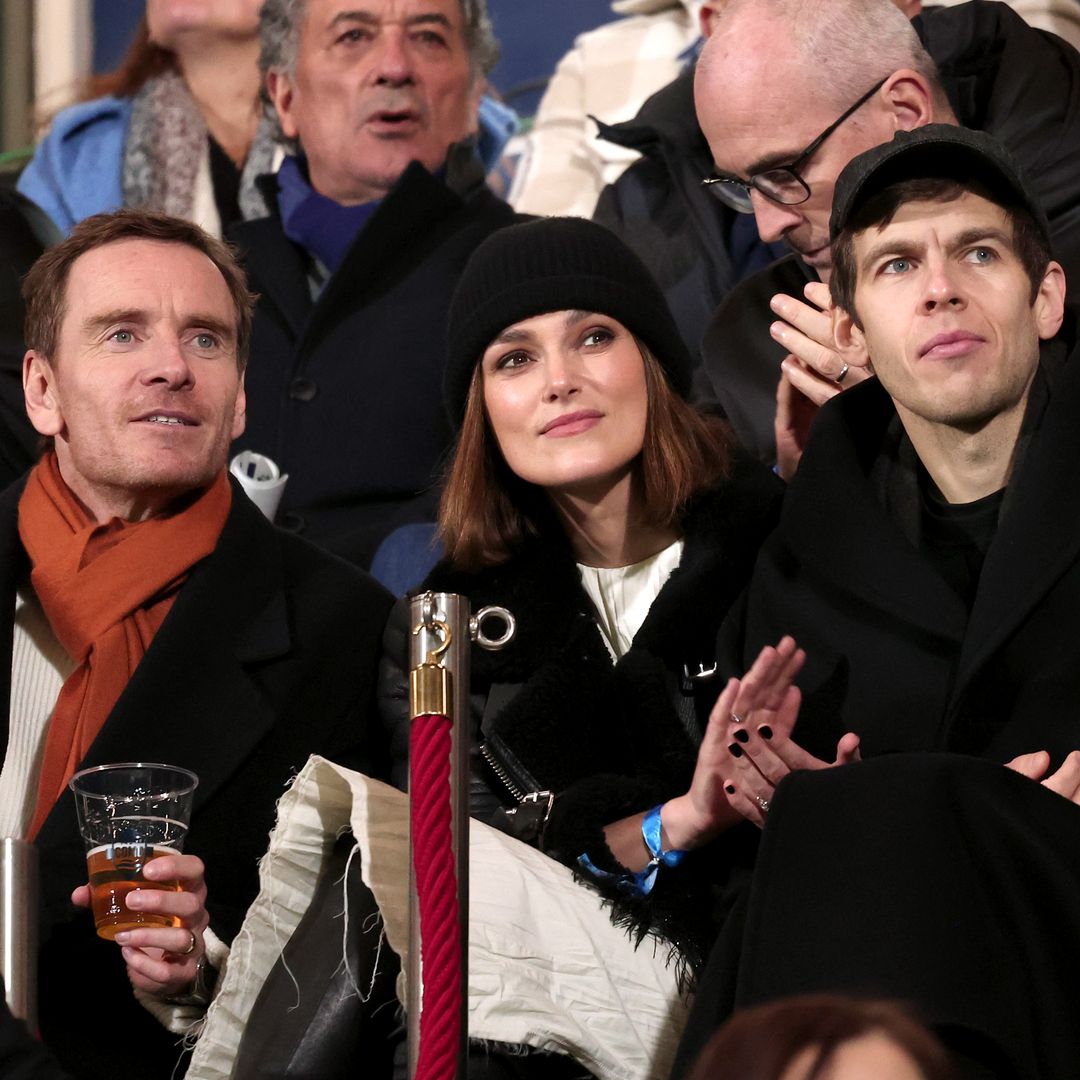  Seated at the stadium, Keira Knightley wears her chic black leather jacket and a casual black beanie. Smiling next to Michael Fassbender and James Righton, she applauds a thrilling match moment, her ensemble blending comfort with couture.