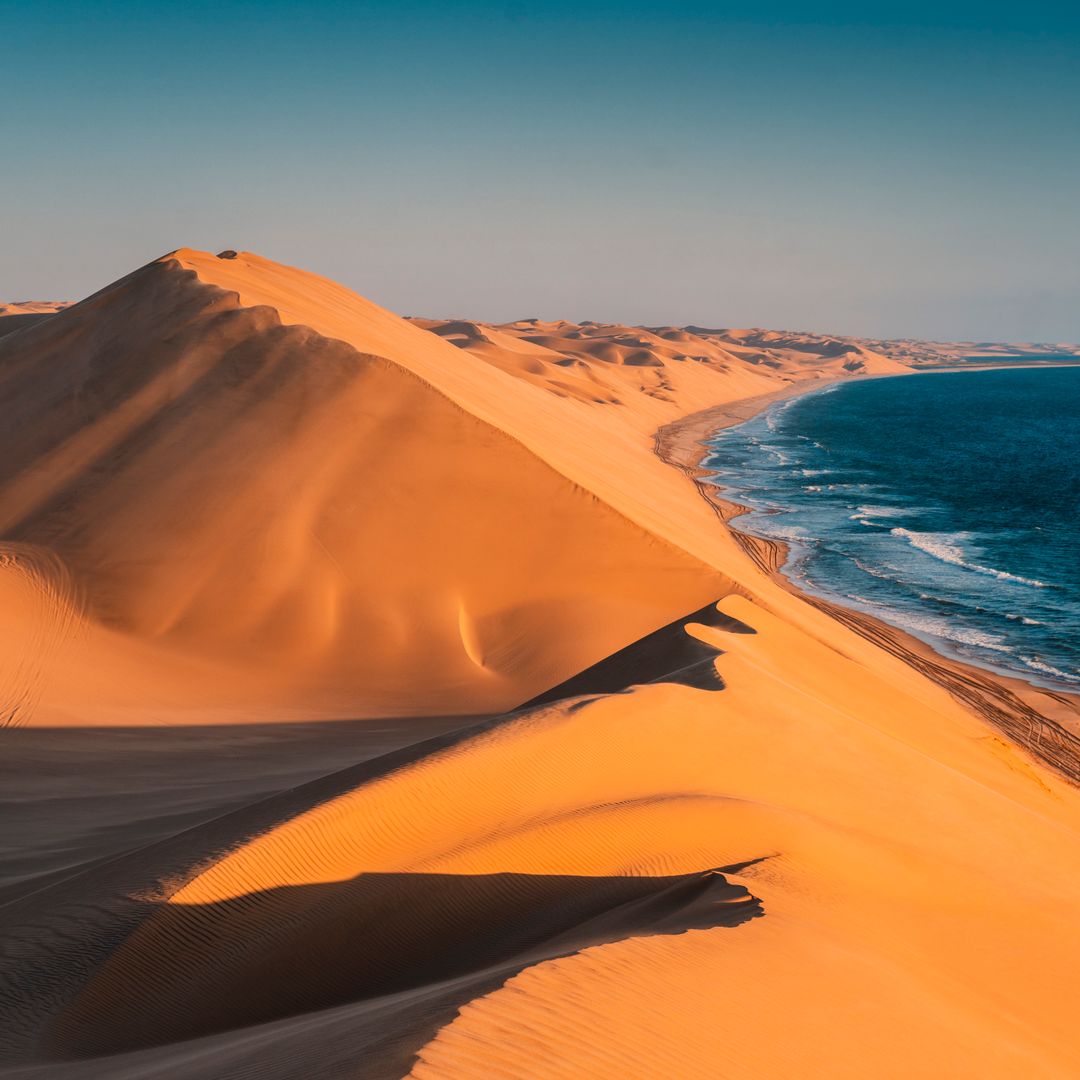 A vast desert landscape meeting the ocean. The golden sand dunes extend towards the deep blue sea, creating a striking contrast