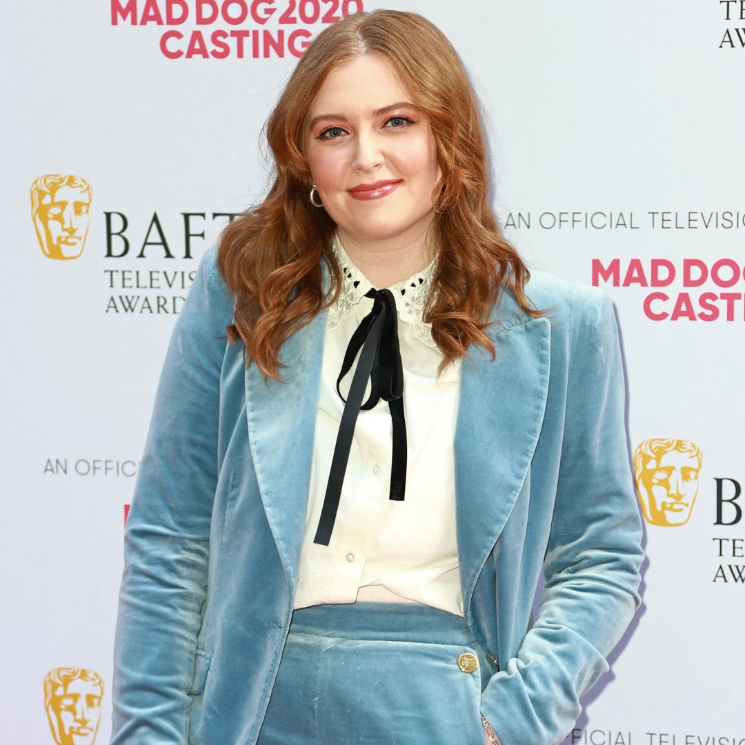 Alice Oseman smiling for a photo in a blue velvet suit on a red carpet