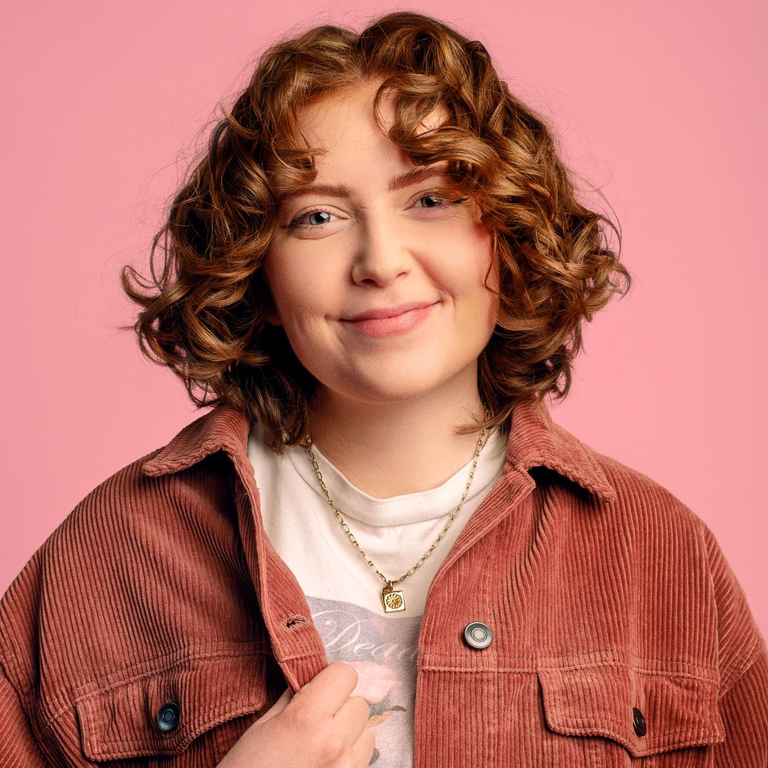 Alice Oseman smiling photographed in front of a pink background