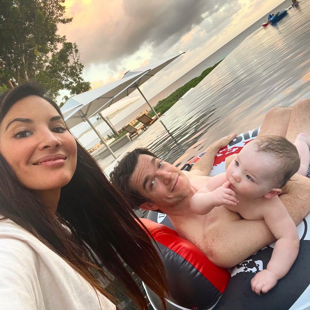 Olivia smiling for a selfie with John and Malcolm in the background floating on a float in a pool