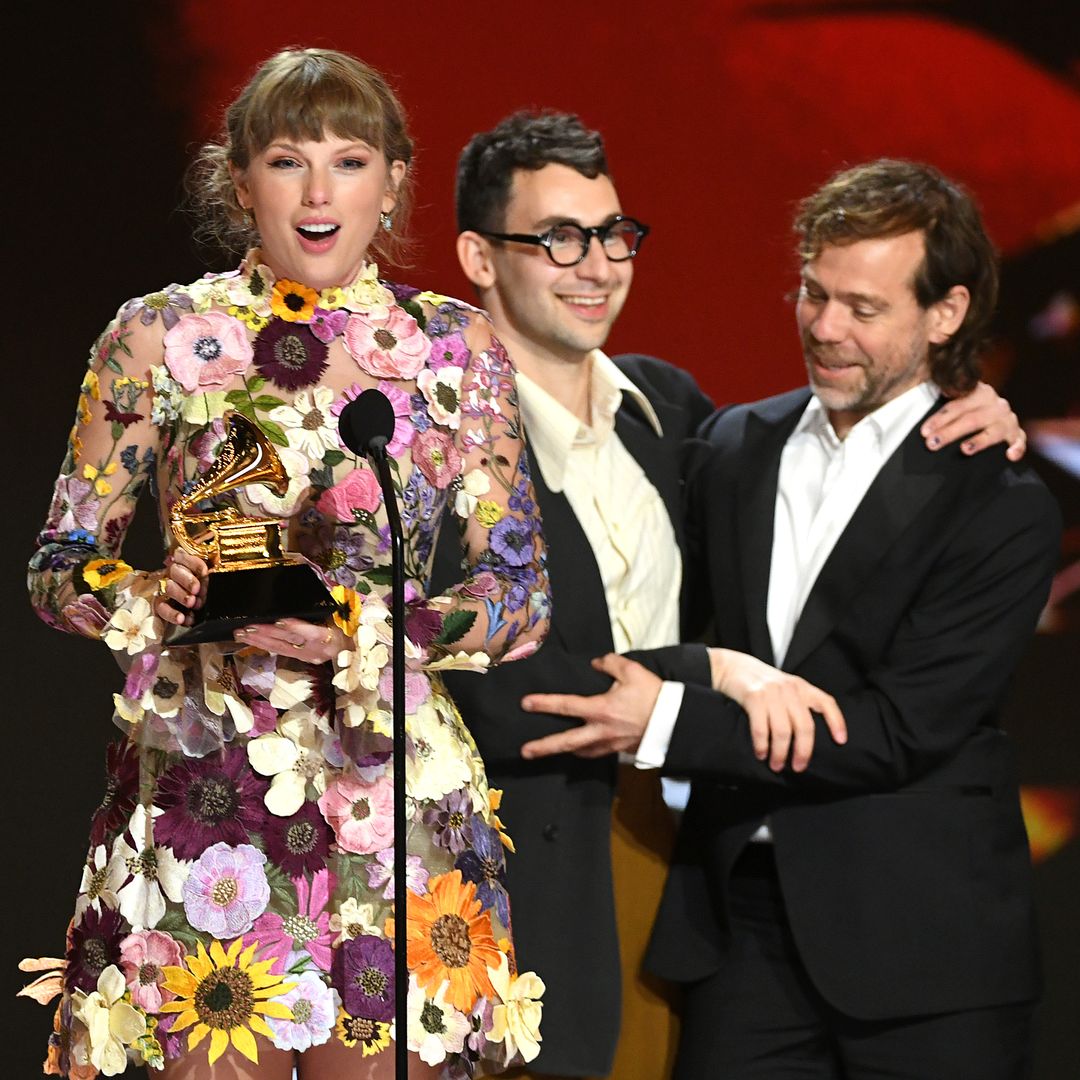 Taylor Swift speaking into a mic while holding a Grammy, her team celebrating behind her