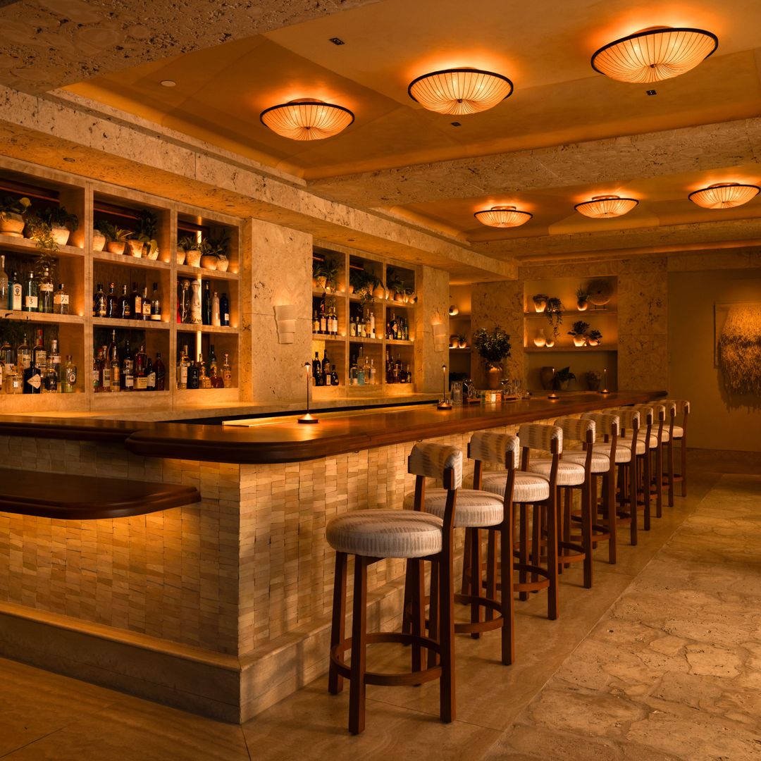 The bar at AVIV restaurant in 1 Hotel South Beach, featuring a long, elegant wooden counter lined with cushioned barstools. Warm, ambient lighting glows from sculptural ceiling fixtures and recessed wall sconces, creating a cozy, intimate atmosphere.