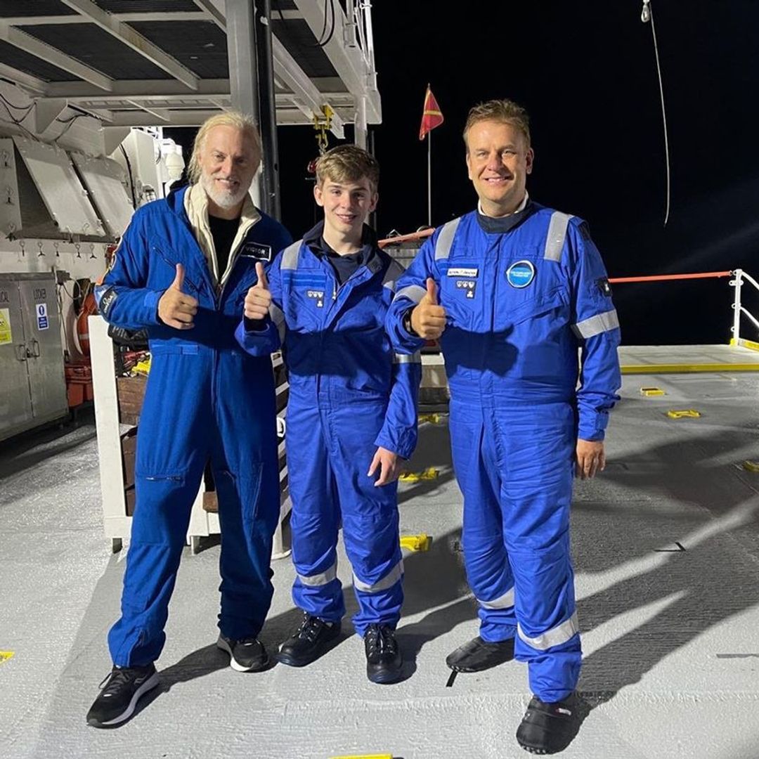 Hamish posing for a photo with his son Giles and fellow undersea explorer Victor Vescovo all are wearing blue boiler suits and smiling with a thumbs up